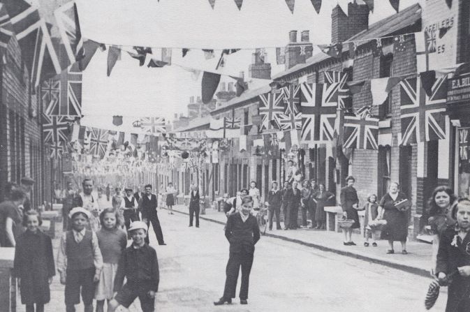 A black and white photo of a group of people having a party in the street. There are Union flags bunting between the houses.